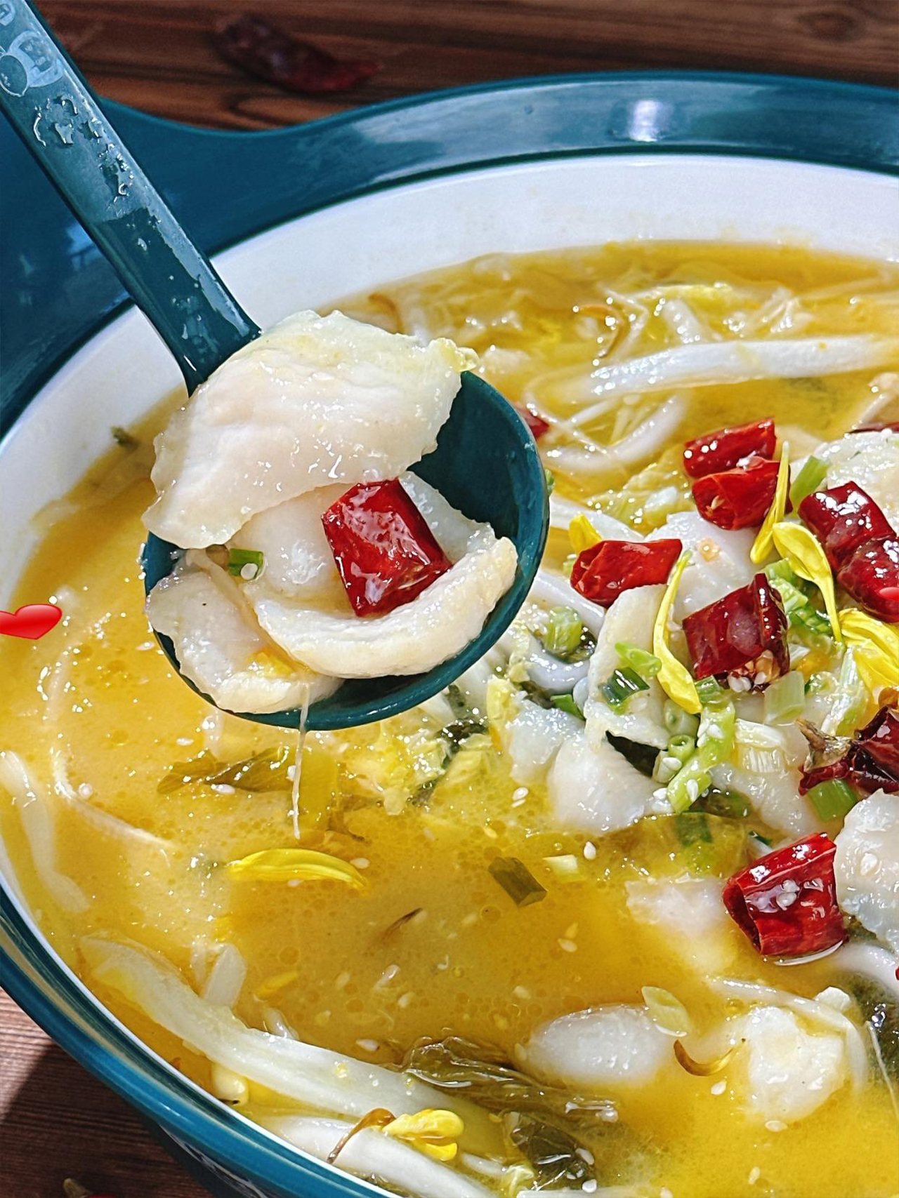 A close-up of a spoon lifting tender white fish fillets from a spicy, yellow broth filled with chili peppers and bean sprouts.
